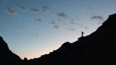 Siluet, karanlık dağ sırasının turuncu mavi silueti, mutlu erkek yürüyüşçü, zirvede, zevkle el kaldırıyor, duygularla boğulmuş. Yüksek kalite 4k görüntü