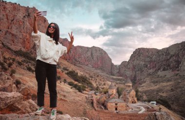 Mutlu kadın turist 13. yüzyıl Ermeni manastırının arka planında renkli kayalıklarla çevrili bir selfie çekiyor.