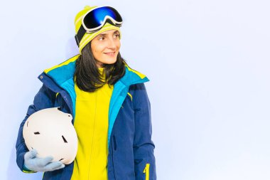 Kış sporları konsepti. Mavi ceketli, kayak gözlüklü, yeşil kazaklı, kasklı, mavi arka planlı güzel bir kadın. Aktif kış tatili hazırlığı ve giyimi