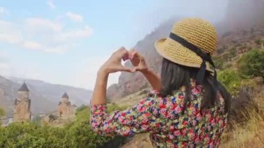 Mutlu genç turist kameraya bakar ve kalp şeklinde poz verir. Renkli gündoğumu sabahında Noravank Manastırı 'nın panorama manzarasına karşı. Aşk dolu kalp şeklinde eller, aşk konsepti.