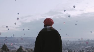 Close up back view slow motion Woman looking around observe,wear fashionable clothing stand on scenic viewpoint watch hot air balloon flight on sunrise with beautiful goreme town and valley background