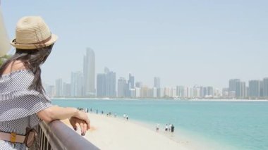 Kadın turist, turist grubuyla birlikte kumlu turkuaz su sahili manzarasına hayran. Abu dhabi Metropolis gökyüzü geçmişi. Gezgin tatilin tadını çıkar emirlikleri ve körfezi keşfet