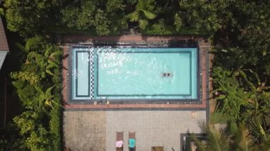 Havadan aşağıya zum yap, tropikal manzarayı izle. Genç bir kadın kristal berrak havuzda huzur içinde yüzüyor. Etrafı gür orman bitkileri ve palmiye ağaçlarıyla çevrili. İyilik ve refah tropikal adalarda geri çekiliyor.