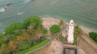 Sri Lankalıların Galle Kalesi 'ndeki ikonik beyaz deniz fenerinin yanında okyanusta yüzüşlerini gösteren dairesel hava manzarası. Sahne, tarihi mimariyi günlük kıyı yaşamı ve ada eğlencesi ile harmanlıyor.