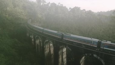Hava manzaralı yolcu treni, bulutlu sabahın erken saatlerinde yemyeşil sularla çevrili Ella, Sri Lanka yakınlarındaki ikonik dokuz kemer köprüsünü geçer.