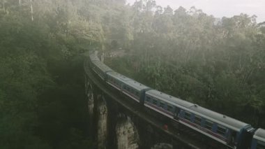 Ağır çekimde uçan yolcu treni bulutlu bir sabah erken saatlerde yemyeşil sularla çevrili Ella, Sri Lanka yakınlarındaki ikonik dokuz kemer köprüsünü geçer.