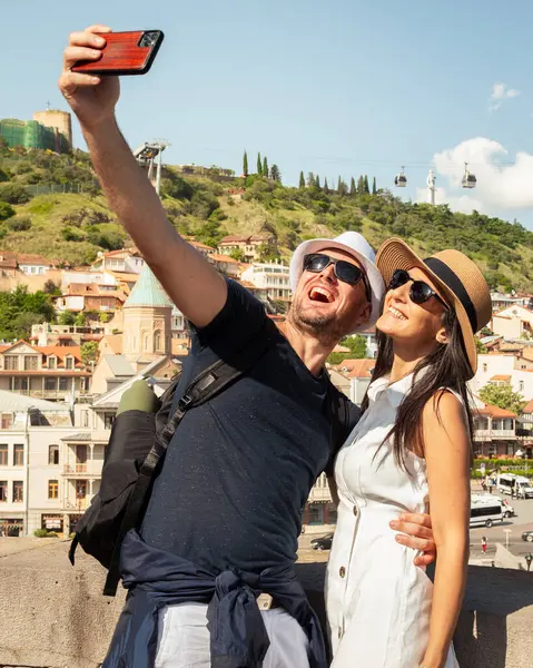 Mutlu çift, arka planda Narikala Kalesi ve teleferiği ile Tiflis Eski Şehir 'in önünde selfie çekiyor. Aşkı, bağlantıyı, seyahat neşesini, modern romantizmi yakalıyorlar..