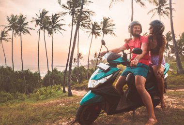 Güneydoğu Asya 'da Sri Lanka' da palmiye ağaçları ve sahil şeridi boyunca motosikletli mutlu beyaz çift tropikal günbatımının tadını çıkarıyor. Yolculuk ve scooter kiralama konsepti