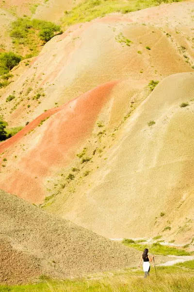 Kot pantolonlu kadın yürüyüşçü, Georgia 'nın Mravaltskaro gökkuşağı dağlarının canlı şeritli tepelerinin üzerinde engin bozkır arka planına sahip çiğ patika yolunda yürüyor. Mravaltskaro Renkli Dağları