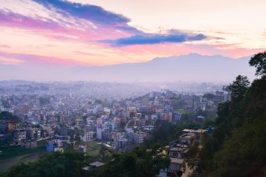 Katmandu, Nepal üzerinde geniş panoramik günbatımı manzarası, yoğun kentsel yayılma ile canlı bir gökyüzü altında ve akşam ışığı altında parlayan soluk Himalaya dağı arkaplanı