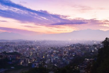Katmandu, Nepal üzerinde geniş panoramik günbatımı manzarası, yoğun kentsel yayılma ile canlı bir gökyüzü altında ve akşam ışığı altında parlayan soluk Himalaya dağı arkaplanı