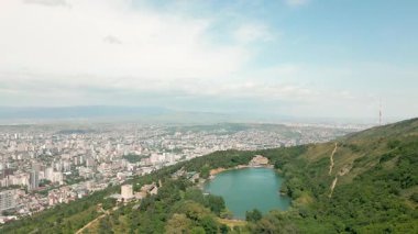Hava manzaralı Kaplumbağa Gölü Tiflis 'in yukarısındaki ormanlık tepelerle çevrili modern şehir manzarası, Mtatsminda sırtı, TV kulesi, yaz havası ve panoramik gökyüzü.