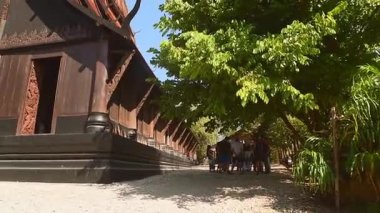 Chiang Rai, Tayland - 2024: Geleneksel ahşap binalar, karmaşık oymalar ve güneş ışığı altında tropikal yeşillikler içeren Kara Ev Müzesini ziyaret eden turist grubu