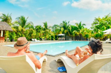 Çiftler güneşli bir günde güneşlenmek için güneşli yataklara uzanır. Tropikal bir tatil köyünde soğuk bir içecek içerler. Yüzme havuzu da güneşli bir tropikal günde Hindistan cevizi palmiyeleriyle çevrilidir.