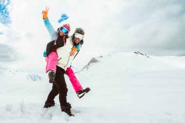 Neşeli genç çift derin karların içinde yürür karlı dağda kameraya bakar, aşkı, mutluluğu, özgürlüğü, eğlenceli ilişkiyi, macerayı, tasasız kış kaçışını, birlikteliği simgeler.