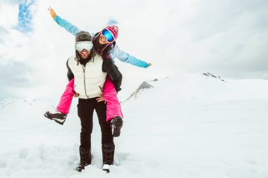 Neşeli genç çift derin karların içinde yürür karlı dağda kameraya bakar, aşkı, mutluluğu, özgürlüğü, eğlenceli ilişkiyi, macerayı, tasasız kış kaçışını, birlikteliği simgeler.