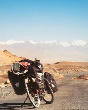 Dört pannier çantasına yüklenmiş kırmızı bisiklet. Çakıl yolunun kenarında duruyor. Etrafı vahşi Pamir dağlarıyla çevrili. Yalnız macera seyahati