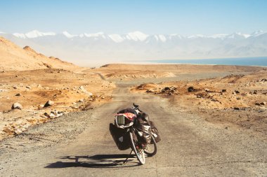 Dört pannier çantasına yüklenmiş kırmızı bisiklet. Çakıl yolunun kenarında duruyor. Etrafı vahşi Pamir dağlarıyla çevrili. Yalnız macera seyahati