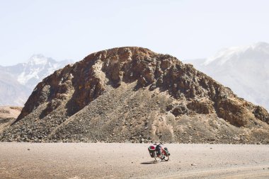 Dört pannier çantasına yüklenmiş kırmızı bisiklet. Çakıl yolunun kenarında duruyor. Etrafı vahşi Pamir dağlarıyla çevrili. Yalnız macera seyahati