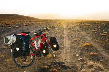 Dört pannier çantasına yüklenmiş kırmızı bisiklet. Çakıl yolunun kenarında duruyor. Etrafı vahşi Pamir dağlarıyla çevrili. Yalnız macera seyahati