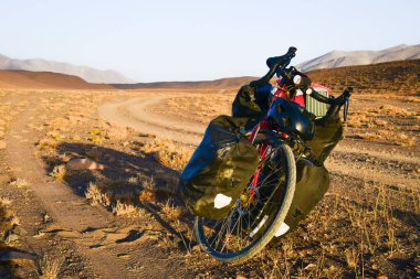 Dört pannier çantasına yüklenmiş kırmızı bisiklet. Çakıl yolunun kenarında duruyor. Etrafı vahşi Pamir dağlarıyla çevrili. Yalnız macera seyahati