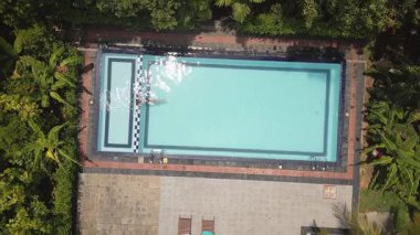 Havadan aşağıya bakan tropik manzara. Genç bir kadın kristal berrak havuzda huzur içinde yüzüyor. Etrafı yemyeşil orman bitkileri ve palmiye ağaçlarıyla çevrili. İyilik ve refah tropikal adalarda geri çekiliyor.