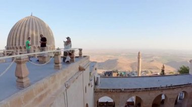 Mardin, Türkiye - 17 Ağustos 2025: Timelapse Zinciriye Medresesi terasını, düğün gruplarını ve taşınan ziyaretçileri, kubbeleri ve minare ışıklarını, Mezopotamya ovasının üzerindeki Eski Mardin ufuk çizgisi alacakaranlıkta.