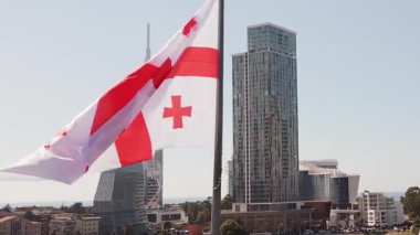 Gökyüzü yakın makro görüntü Gürcistan ulusal bayrağı ve Batumi şehir bayrağı dalgalanan şehir modern siluet mimarisi, güneşli sahil kıyısı ve Karadeniz kıyısı simgeleri ile bayrak direkleri üzerinde