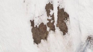 Eriyen karla kaplı kayak yamacının üzerinde uçan hava, iklim değişikliğini, küresel ısınmayı ve kış mevsimindeki düşüşü simgeleyen büyük kahverengi toprak parçalarını ve rayları ortaya çıkarıyor. Kayak sezonunun başı ya da sonu