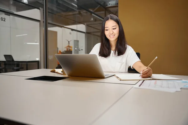 Front view portrait of smiling Asian female analyst looking to the screen of laptop while taking notes in the sketchbook in modern conference room