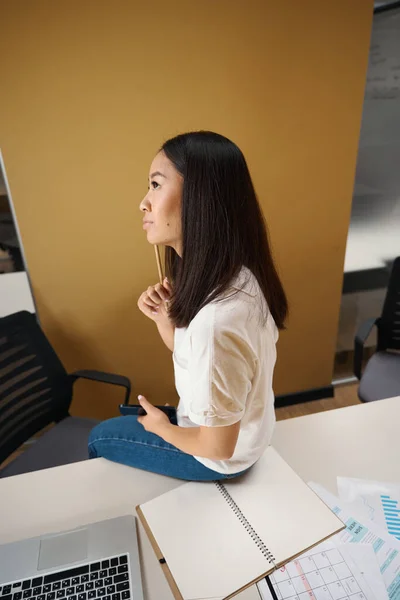 明るいオフィスのオフィスデスクに座っている間に手に鉛筆で思慮深い女性のリアビューの肖像画 — ストック写真