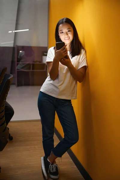 オフィスの明るい廊下には スマートフォンを手にしたエレガントなアジアのオフィスワーカーの完全な長さの肖像画が立っています — ストック写真