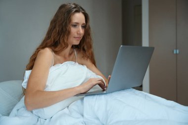 Odaklanmış kadın, dizüstü bilgisayar kullanarak yatakta otururken online sorunları çözmekle meşgul.