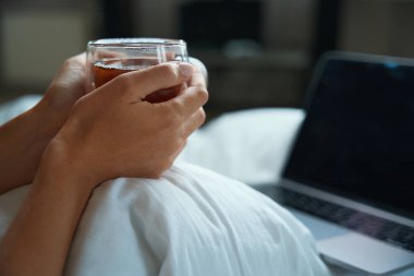 Düzgün manikürlü bir kadın elleri sıcak bir içecekle ısıtılır ve yakınlarda açık bir laptop bulunur.
