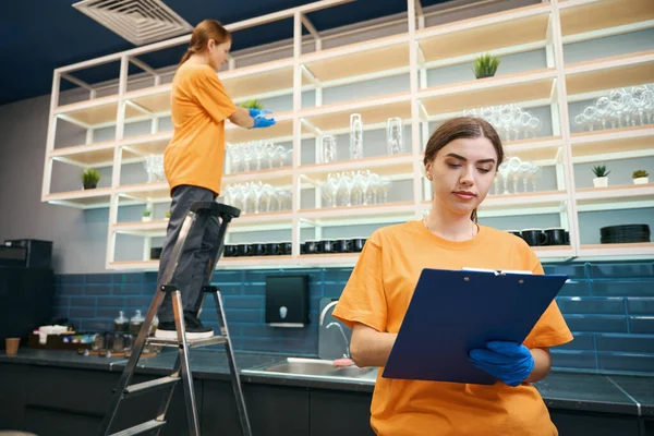 Cleaning company workers checking the list of cleaned areas in a coworking space, women in yellow uniforms and protective gloves