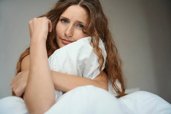 Pensive lady sits on the bed while holding the edge of the blanket with her hands