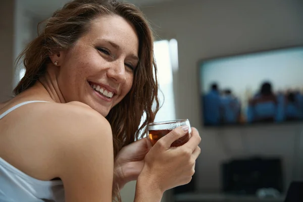 Joyful lady with bare shoulders and flowing hair holds a cup of tea while a wide smile is on her face