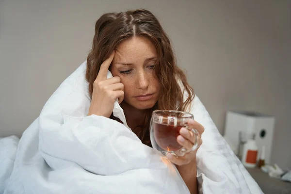 一位疲惫不堪的女士在家里的床上挣扎着与疾病作斗争 而药物和感冒药则躺在她旁边的床头柜上 — 图库照片