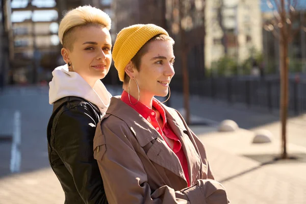 Stock image Smiling blonde in leather jacket hugged her friend in beige trench coat from back, they are LGBT couple on awalk