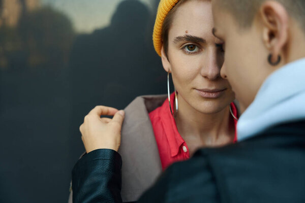 LGBT female couple in modern clothes stands against background of gray wall, woman put her hand on shoulder of friend