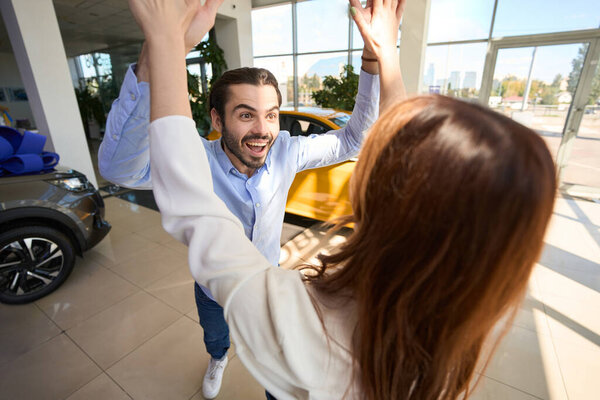 Joyful guy giving high five to his female companion at auto dealership