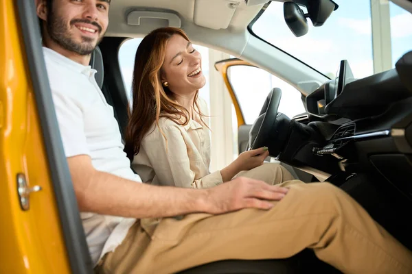 Joyful female driver sitting behind steering wheel next to pleased young man