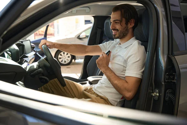 Joyful guy with car key fob sitting in automobile and turning steering wheel