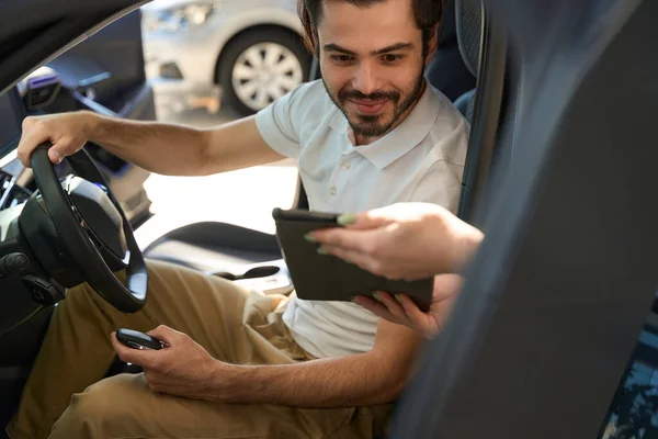 Pleased auto dealership client sitting inside new automobile and looking at tablet computer in salesperson hands