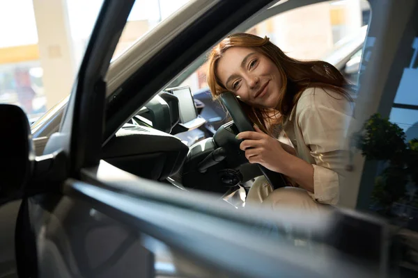 Smiling contented young automotive dealership customer embracing steering wheel of her vehicle