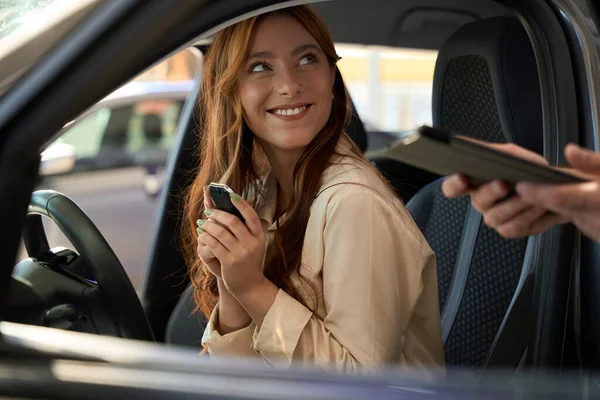 Happy lady with auto key fob sitting at steering wheel and looking at salesperson with tablet computer in hands