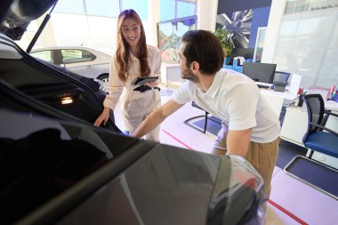 Gülümseyen genç otomobil satıcısı bagaj bölümünü potansiyel alıcıya gösteriyor.