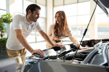 Odaklanmış otomotiv alıcısı satış elemanının huzurunda araba kaputunun altındaki parçalara bakıyor.