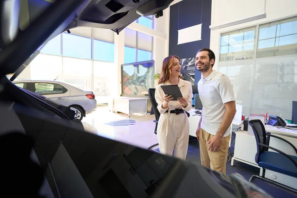 Joyous car salesperson with tablet computer in hand looking at pleased buyer