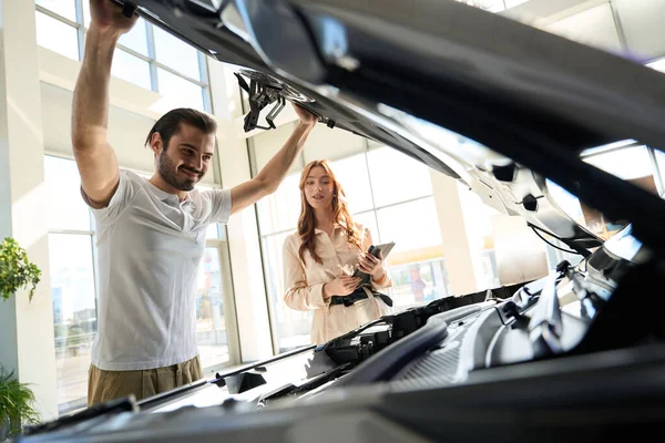 営業女性の前でオープンカーのボンネットの下に自動車部品を検討笑みを浮かべて幸せな買い手 — ストック写真